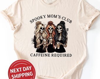 May include: Cream-colored t-shirt featuring a graphic of three skeleton women holding coffee cups. The text "Spooky Mom's Club Caffeine Required" is above and below the image. The shirt has short sleeves and a round neckline.