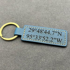 May include: A blue leather keychain with a gold-colored key ring. The rectangular tag has stitched edges and displays the coordinates 29°48'44.7"N 95°33'52.2"W in black text. The keychain rests on a gray surface.
