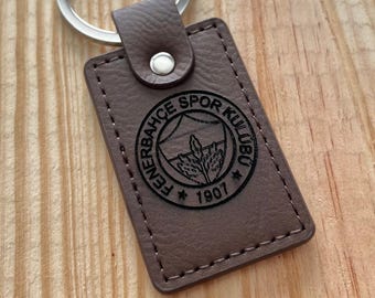 May include: A brown leather keychain with a silver ring. The rectangular tag features the Fenerbahce Spor Kulubu logo and the year 1907. The keychain is on a wooden surface.