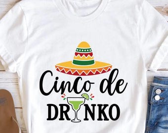 May include: White t-shirt with a colorful sombrero graphic and the text "Cinco de DRINKO". The design includes a margarita glass. The shirt is paired with denim shorts and sandals, suggesting a casual, festive theme.