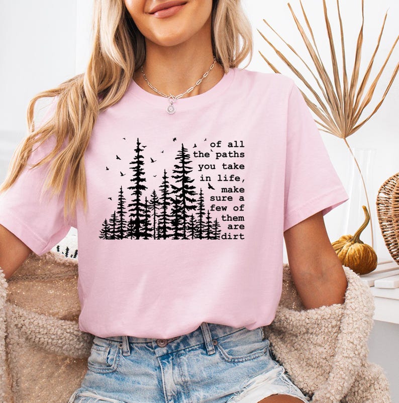 May include: A light pink short-sleeve t-shirt featuring a black graphic of a forest with birds and the text: of all the paths you take in life, make sure a few of them are dirt. The shirt has a crew neckline and a standard fit design.