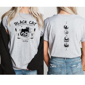 May include: A heather gray t-shirt featuring a black cat on a pumpkin with the text "Black Cat Mystical Brews Fine Potions Apothecary". The back of the shirt displays a vertical design of moon phases, crystals, and an open book.