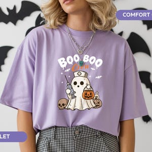 May include: Violet-colored t-shirt with a Halloween-themed graphic. The design features a ghost wearing a nurse's cap and holding a syringe and a jack-o'-lantern. The text "BOO CREW" is above the ghost. Black bat cutouts decorate the background.