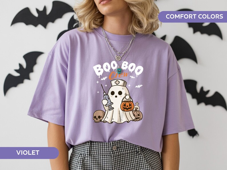May include: Violet-colored t-shirt with a Halloween-themed graphic. The design features a ghost wearing a nurse's cap and holding a syringe and a jack-o'-lantern. The text "BOO CREW" is above the ghost. Black bat cutouts decorate the background.
