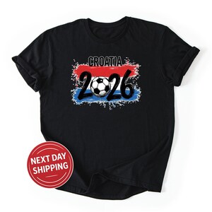 May include: Black t-shirt with a graphic featuring the word "CROATIA" above the number "2026" and a soccer ball. The design incorporates red, white, and blue colors, resembling a flag. A red and white "NEXT DAY SHIPPING" sticker is on the shirt.