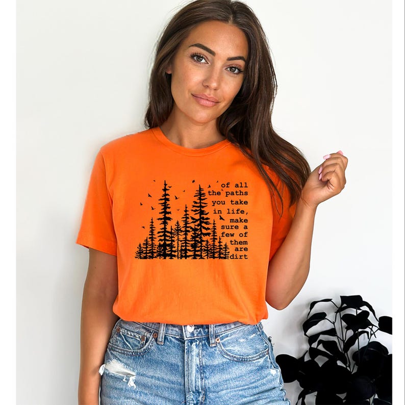 May include: A bright orange short-sleeved t-shirt featuring a black graphic print of a forest silhouette with birds and the text: Of all the paths you take in life, make sure a few of them are dirt. The shirt has a crew neckline and a standard fit.