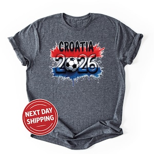 Puede incluir: Camiseta gris oscuro con el texto "CROATIA 2026" y un dise&ntilde;o gr&aacute;fico de bal&oacute;n de f&uacute;tbol. El dise&ntilde;o incorpora los colores rojo, blanco y azul, con un efecto de pintura goteando. La camiseta tiene mangas enrolladas.