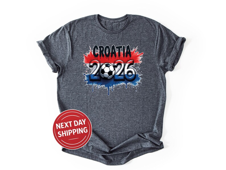 Puede incluir: Camiseta gris oscuro con el texto "CROATIA 2026" y un dise&ntilde;o gr&aacute;fico de bal&oacute;n de f&uacute;tbol. El dise&ntilde;o incorpora los colores rojo, blanco y azul, con un efecto de pintura goteando. La camiseta tiene mangas enrolladas.