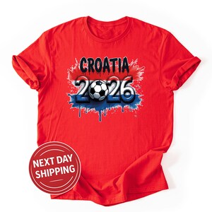 Puede incluir: Camiseta roja con la palabra "CROATIA" y el n&uacute;mero "2026" con un bal&oacute;n de f&uacute;tbol. El dise&ntilde;o utiliza los colores rojo, blanco y azul con un efecto de pintura goteando. La camiseta es de cuello redondo.