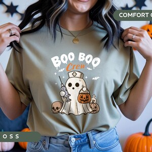 May include: Moss green t-shirt with a Halloween-themed graphic. The design features a ghost wearing a nurse's cap, holding a syringe and a jack-o'-lantern. The text "BOO BOO Crew" is above the ghost. Comfort Colors.