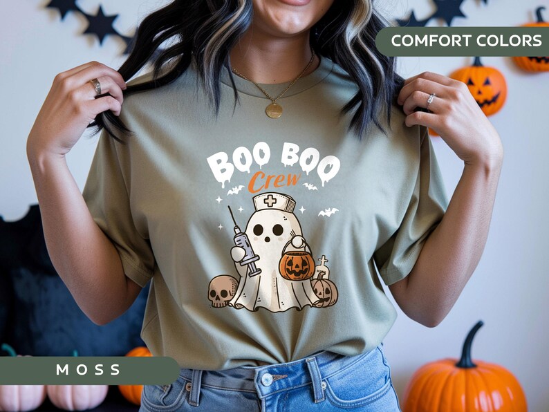 May include: Moss green t-shirt with a Halloween-themed graphic. The design features a ghost wearing a nurse's cap, holding a syringe and a jack-o'-lantern. The text "BOO BOO Crew" is above the ghost. Comfort Colors.