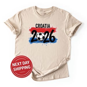 May include: Beige t-shirt with the word "CROATIA" in black above a red, white, and blue design. Below the design is the number "2026" in black, with a soccer ball in the center. A red circle with "NEXT DAY SHIPPING" is in the lower left.