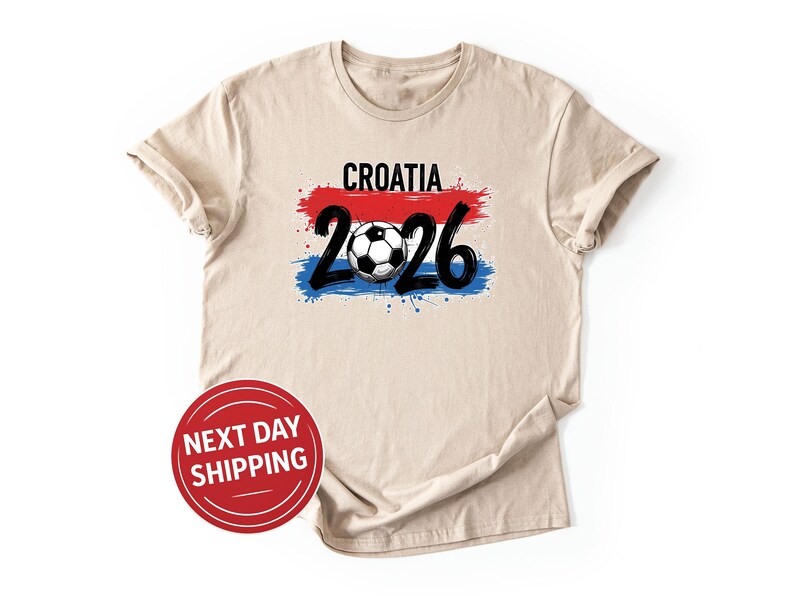 May include: Beige t-shirt with the word "CROATIA" in black above a red, white, and blue design. Below the design is the number "2026" in black, with a soccer ball in the center. A red circle with "NEXT DAY SHIPPING" is in the lower left.
