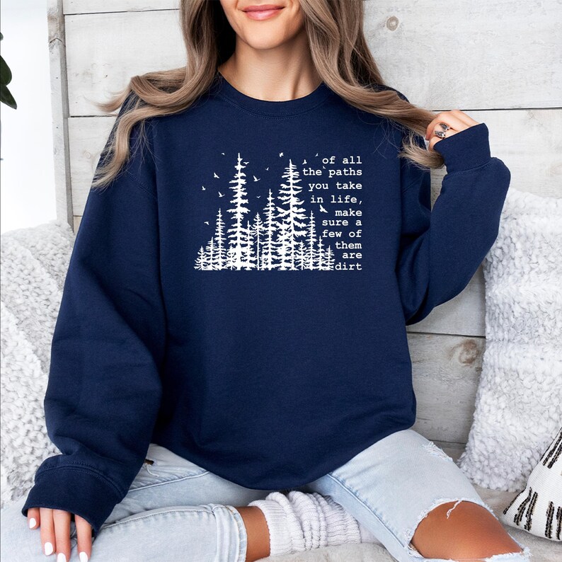 May include: A navy blue crewneck sweatshirt featuring a white graphic print of a forest of pine trees and birds. The text reads: of all the paths you take in life, make sure a few of them are dirt. The garment has long sleeves and ribbed cuffs.