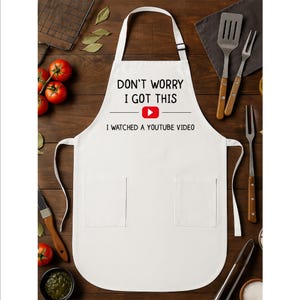 May include: White apron with the text "DON'T WORRY I GOT THIS" and "I WATCHED A YOUTUBE VIDEO" in black font. The YouTube logo is in the center. The apron has two pockets and a neck strap. Kitchen utensils and tomatoes are in the background.