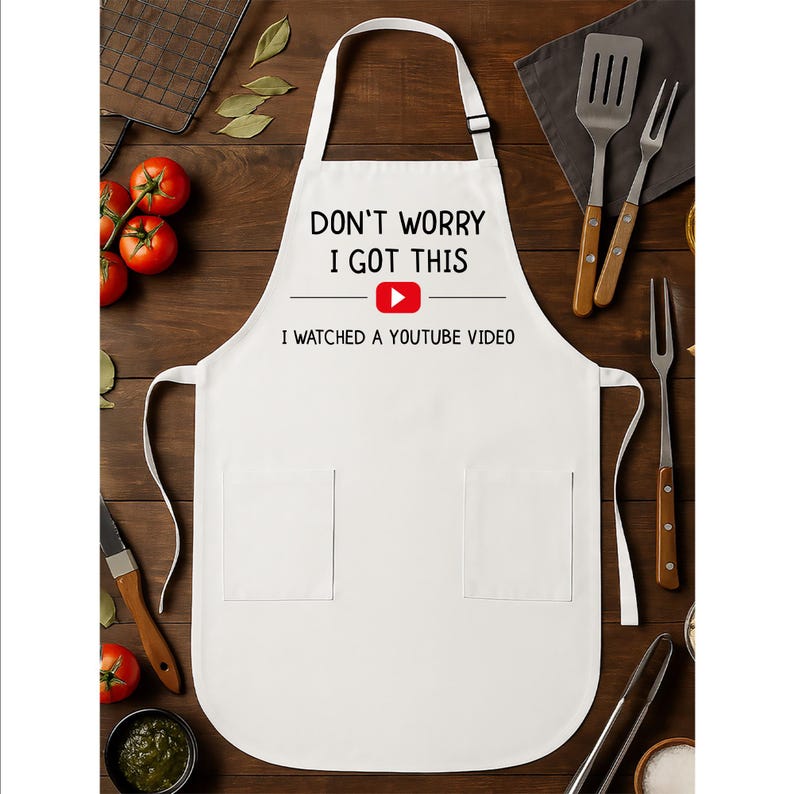 May include: White apron with the text "DON'T WORRY I GOT THIS" and "I WATCHED A YOUTUBE VIDEO" in black font. The YouTube logo is in the center. The apron has two pockets and a neck strap. Kitchen utensils and tomatoes are in the background.