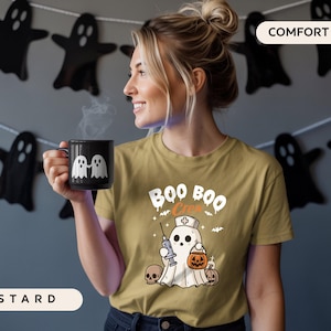 May include: Mustard-colored t-shirt with a Halloween-themed graphic that says "Boo Boo Crew". The design features a ghost dressed as a nurse holding a syringe and a pumpkin. A black mug with two white ghost figures is held by a person.