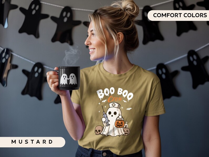May include: Mustard-colored t-shirt with a Halloween-themed graphic that says "Boo Boo Crew". The design features a ghost dressed as a nurse holding a syringe and a pumpkin. A black mug with two white ghost figures is held by a person.