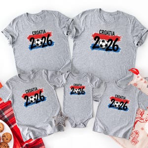 May include: A collection of gray t-shirts and a onesie featuring the text "CROATIA 2026" with a soccer ball graphic. The design includes red, white, and blue accents, suggesting a national theme. The shirts are arranged on a white surface.