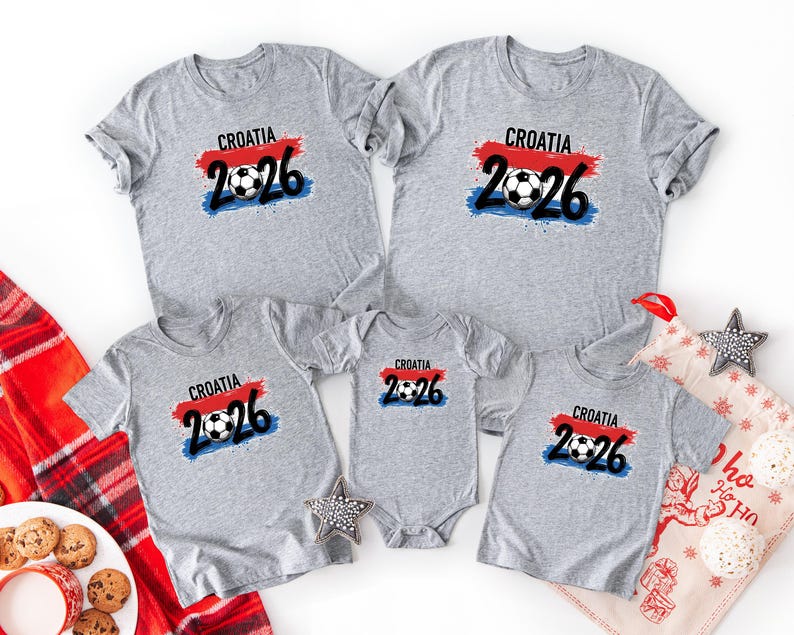 May include: A collection of gray t-shirts and a onesie featuring the text "CROATIA 2026" with a soccer ball graphic. The design includes red, white, and blue accents, suggesting a national theme. The shirts are arranged on a white surface.