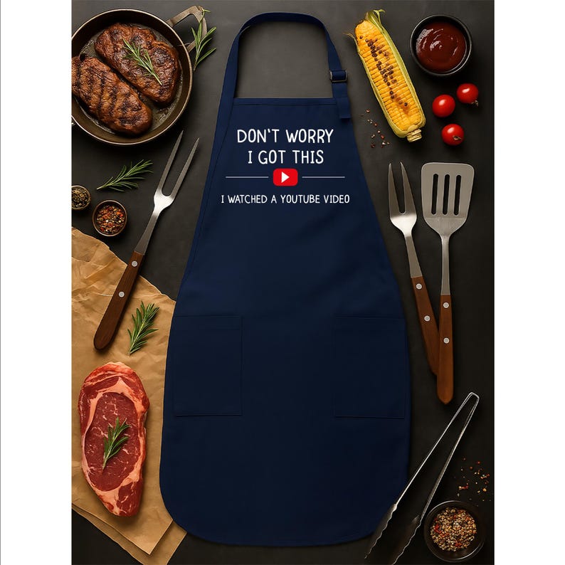 May include: Navy blue apron with the text "DON'T WORRY I GOT THIS" and "I WATCHED A YOUTUBE VIDEO" in white. The apron has two pockets and is surrounded by cooking utensils, steak, and vegetables.