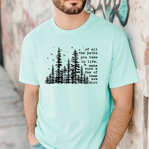 May include: A light mint green short-sleeved t-shirt featuring a black graphic of a forest with birds and the text: of all the paths you take in life, make sure a few of them are dirt. The design is centered on the chest of the garment.