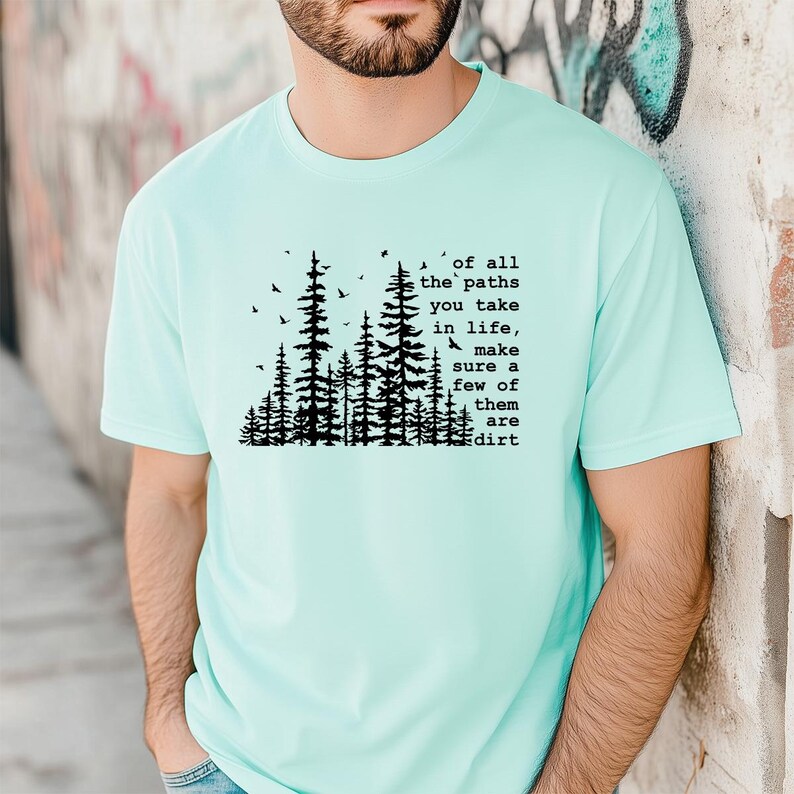 May include: A light mint green short-sleeved t-shirt featuring a black graphic of a forest with birds and the text: of all the paths you take in life, make sure a few of them are dirt. The design is centered on the chest of the garment.