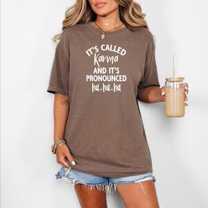 Op de afbeelding: Een bruin T-shirt met de witte tekst "It's Called Karma And It's Pronounced ha-ha-ha". Het shirt wordt gedragen met een spijkerbroek. Een glas met een rietje wordt in de rechterhand gehouden.