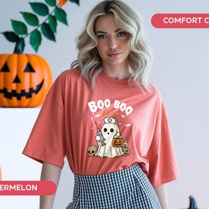 May include: A coral-colored t-shirt with a Halloween-themed graphic. The design features a ghost wearing a nurse's hat, holding a syringe and a pumpkin-shaped treat bag. The words "BOO BOO" are above the ghost. Two carved pumpkins are in the background.