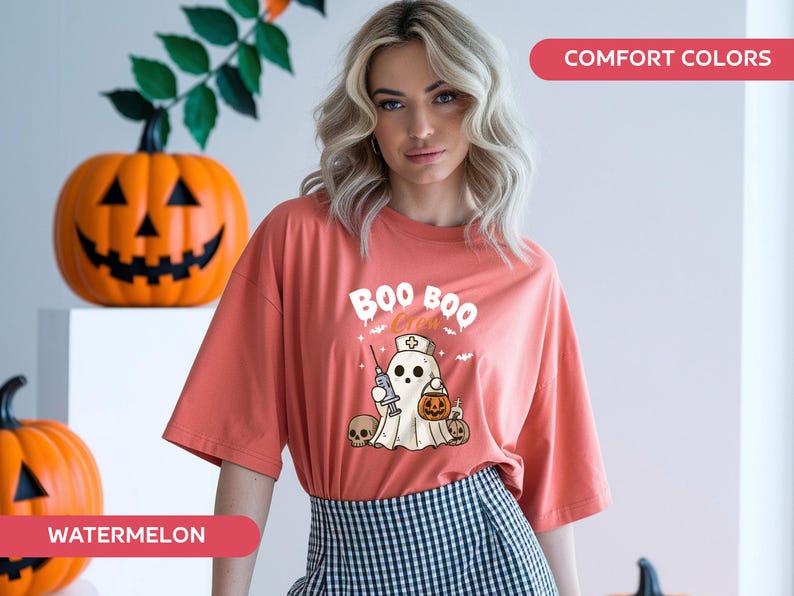 May include: A coral-colored t-shirt with a Halloween-themed graphic. The design features a ghost wearing a nurse's hat, holding a syringe and a pumpkin-shaped treat bag. The words "BOO BOO" are above the ghost. Two carved pumpkins are in the background.
