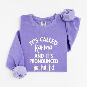 Op de afbeelding: Een lavendelkleurige Comfort Colors sweatshirt met de witte tekst "IT'S CALLED Karma AND IT'S PRONOUNCED ha-ha-ha". De sweatshirt heeft een ronde hals en lange mouwen met manchetten. Het kledingstuk is netjes opgevouwen.