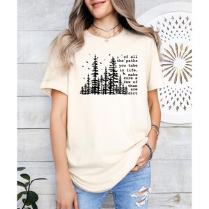 May include: A cream-colored short-sleeve t-shirt featuring a black graphic of a pine forest and the text: of all the paths you take in life, make sure a few of them are dirt. The shirt has a crew neckline and a relaxed fit, worn with blue jeans.
