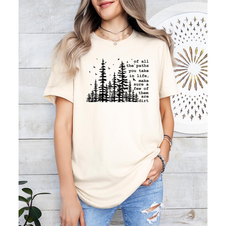 May include: A cream-colored short-sleeve t-shirt featuring a black graphic of a pine forest and the text: of all the paths you take in life, make sure a few of them are dirt. The shirt has a crew neckline and a relaxed fit, worn with blue jeans.