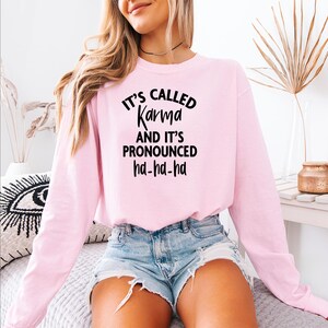 Op de afbeelding: Een lichtroze sweatshirt met de zwarte tekst "It's Called Karma And It's Pronounced ha-ha-ha". De top met lange mouwen wordt gecombineerd met een spijkerbroek. De sweatshirt is een casual, comfortabele stijl.