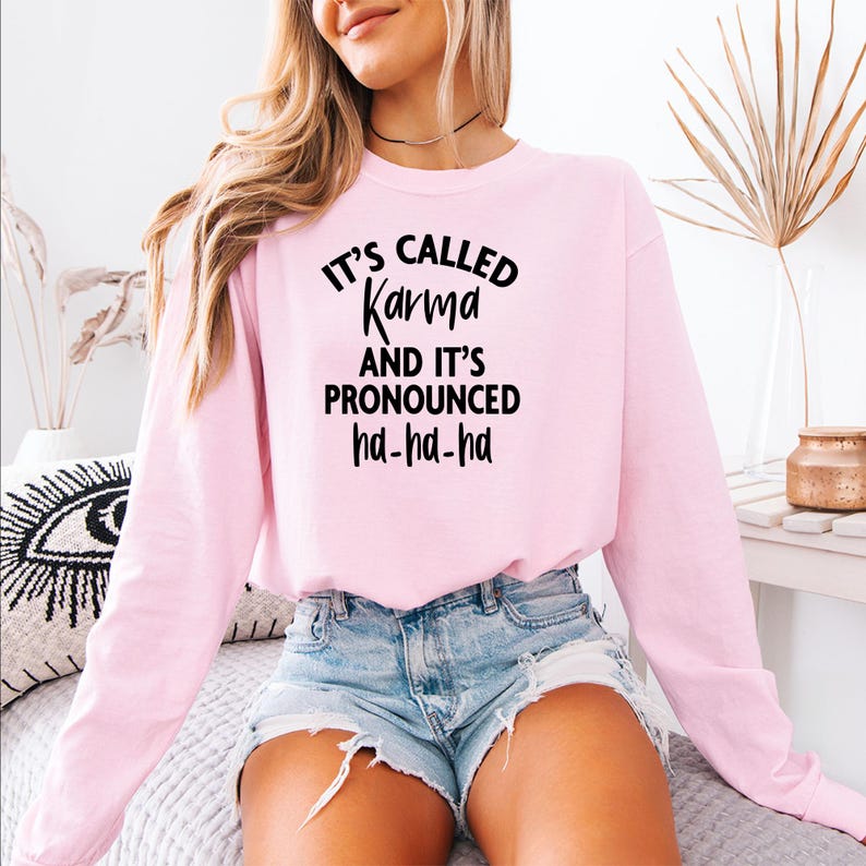 Op de afbeelding: Een lichtroze sweatshirt met de zwarte tekst "It's Called Karma And It's Pronounced ha-ha-ha". De top met lange mouwen wordt gecombineerd met een spijkerbroek. De sweatshirt is een casual, comfortabele stijl.