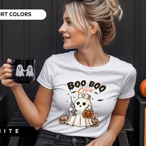 May include: White t-shirt with a Halloween-themed graphic. The design features a ghost wearing a nurse's cap, holding a syringe and a jack-o'-lantern, with the text "Boo Boo Crew". A black mug with two ghost figures is also visible.
