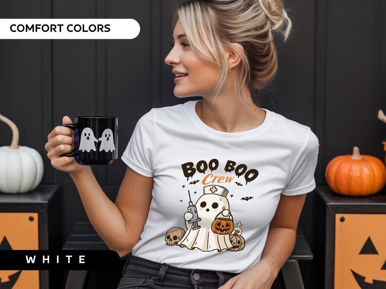 May include: White t-shirt with a Halloween-themed graphic. The design features a ghost wearing a nurse's cap, holding a syringe and a jack-o'-lantern, with the text "Boo Boo Crew". A black mug with two ghost figures is also visible.