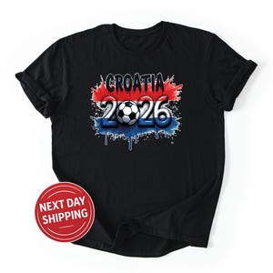 Puede incluir: Camiseta negra con un dise&ntilde;o gr&aacute;fico que presenta la palabra "CROATIA" en blanco, el n&uacute;mero "2026" y un bal&oacute;n de f&uacute;tbol. El dise&ntilde;o incorpora los colores rojo, blanco y azul, con un efecto de pintura goteando. Un c&iacute;rculo rojo con "NEXT DAY SHIPPING" est&aacute; en la parte inferior izquierda.