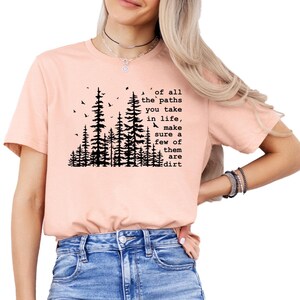 May include: A peach-colored short-sleeve t-shirt featuring a black graphic of pine trees and birds. The text reads: of all the paths you take in life, make sure a few of them are dirt. The shirt is paired with blue denim jeans.