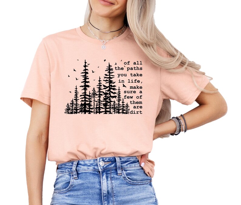 May include: A peach-colored short-sleeve t-shirt featuring a black graphic of pine trees and birds. The text reads: of all the paths you take in life, make sure a few of them are dirt. The shirt is paired with blue denim jeans.