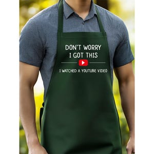 May include: A dark green apron with white text that reads "DON'T WORRY I GOT THIS" above a YouTube play button and the text "I WATCHED A YOUTUBE VIDEO". The apron has a neck strap and waist ties.