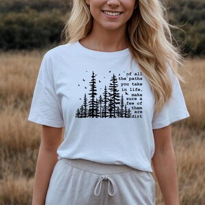 May include: A white short-sleeved t-shirt featuring a black graphic print of a forest silhouette with birds and the text: of all the paths you take in life, make sure a few of them are dirt. The shirt has a crew neckline and a relaxed fit.