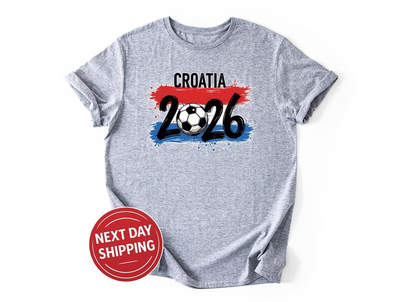 May include: A heather gray t-shirt with the word "CROATIA" above the numbers "2026" and a soccer ball graphic. The design incorporates red, white, and blue brushstrokes, resembling a flag. The shirt is a casual wear item.