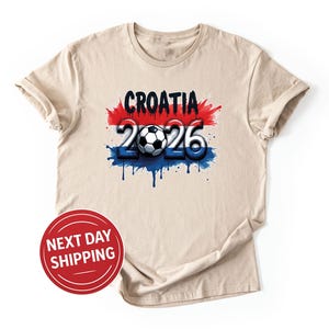 Puede incluir: Camiseta beige con un dise&ntilde;o gr&aacute;fico con la palabra "CROATIA" en negro, el n&uacute;mero "26" en azul y un bal&oacute;n de f&uacute;tbol. El dise&ntilde;o incorpora salpicaduras de pintura roja y azul, y la camiseta tiene una pegatina de "NEXT DAY SHIPPING".