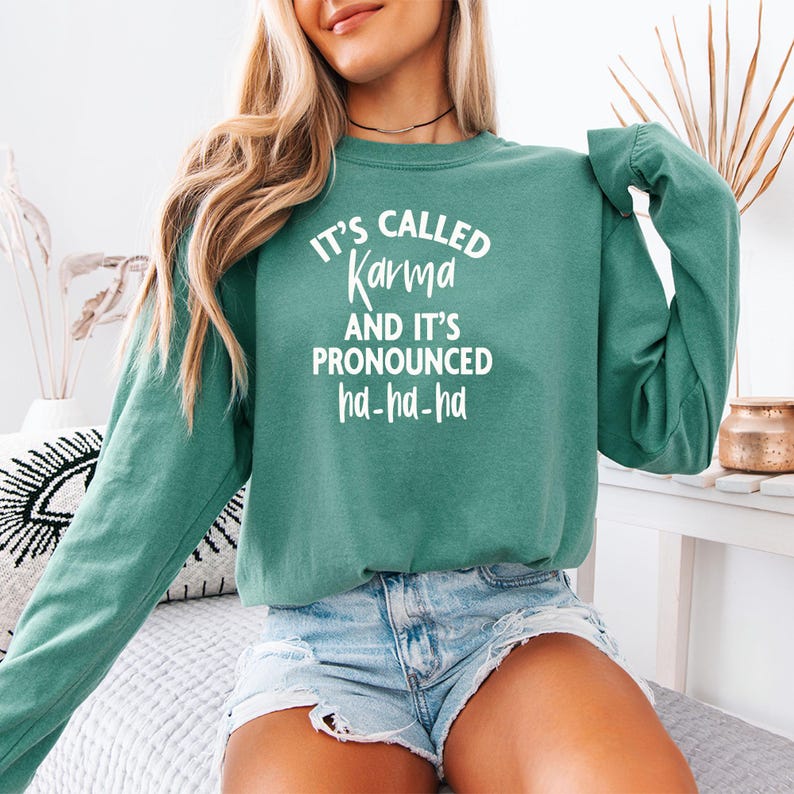 Op de afbeelding: Petrolgroene sweatshirt met de witte tekst "IT'S CALLED Karma AND IT'S PRONOUNCED ha-ha-ha". De top met lange mouwen wordt gecombineerd met een spijkerbroek. De sweatshirt is een casual, comfortabel kledingstuk.