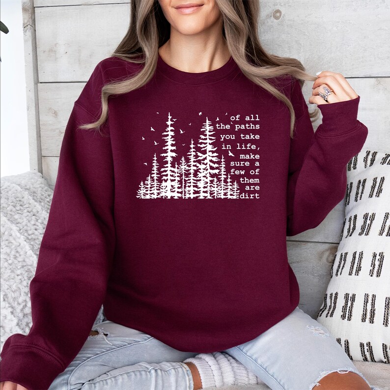 May include: A maroon long-sleeve crewneck sweatshirt featuring a white graphic of pine trees and birds. The text reads: of all the paths you take in life, make sure a few of them are dirt. The design is centered on the chest of the garment.
