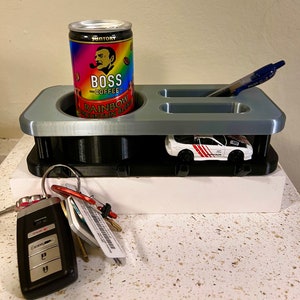 Puede incluir: Un organizador gris impreso en 3D con un portavasos, un porta bolígrafos, un modelo de coche pequeño y ganchos para llaves. El organizador está sobre una superficie blanca.