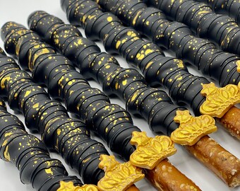 May include: Several chocolate-covered pretzel rods, black with gold flecks, and topped with gold crowns. The pretzels are arranged diagonally against a white background. A tasty treat.