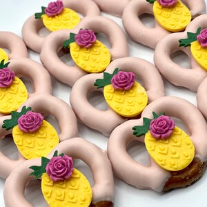 May include: Pink-frosted pretzels, half-dipped in chocolate, are topped with yellow pineapple and purple rose decorations. The treats are arranged on a white surface, creating a visually appealing display of sweet snacks.