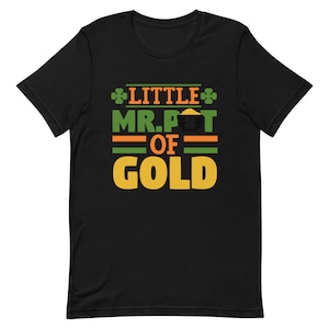 Puede incluir: Camiseta negra con un trébol verde y texto dorado que dice "Little Mr. Pot of Gold".